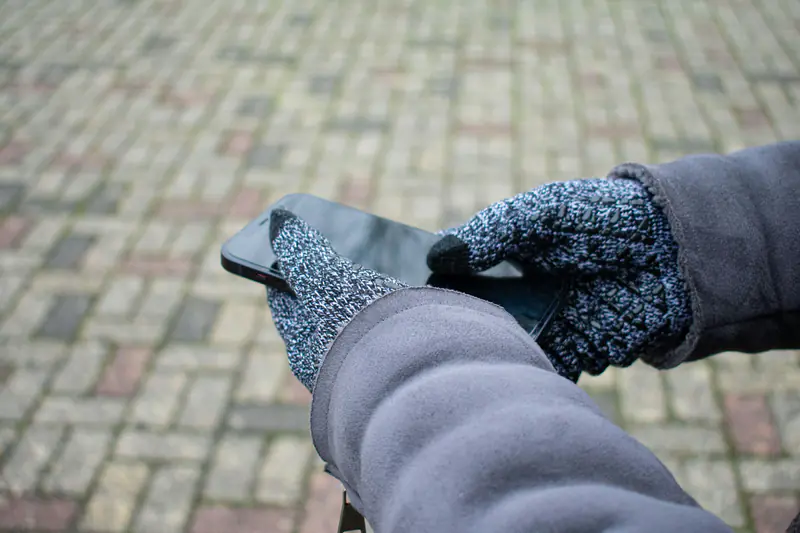 Two hands in grey knit gloves hold a smartphone above a brick paved street