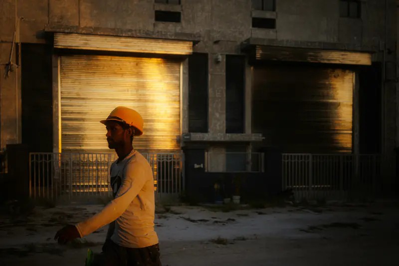 A construction worker in a yellow hard hat walks past a shuttered building in warm evening light