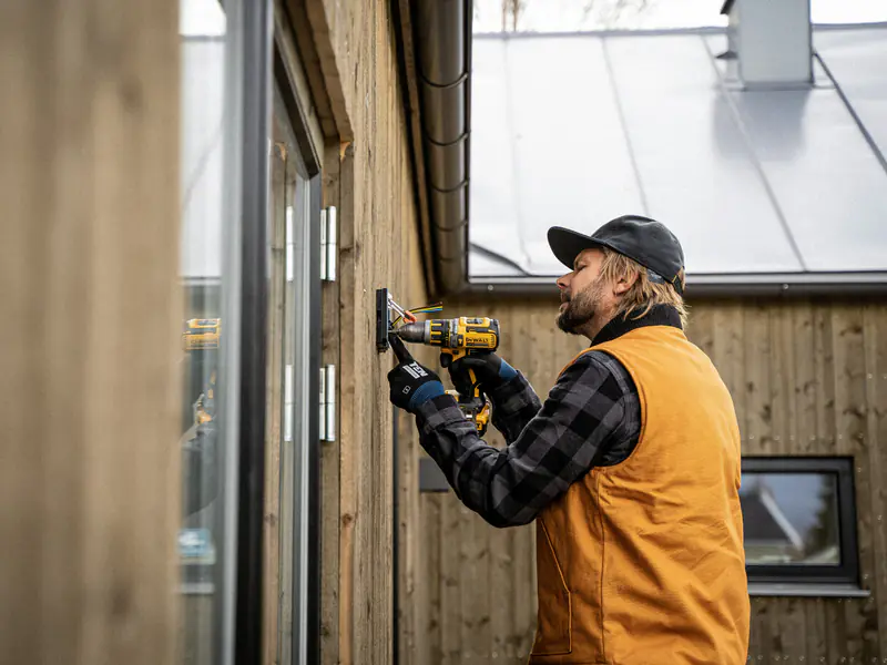 Un artisan en casquette et gilet de travail perce le bardage en bois d’une maison